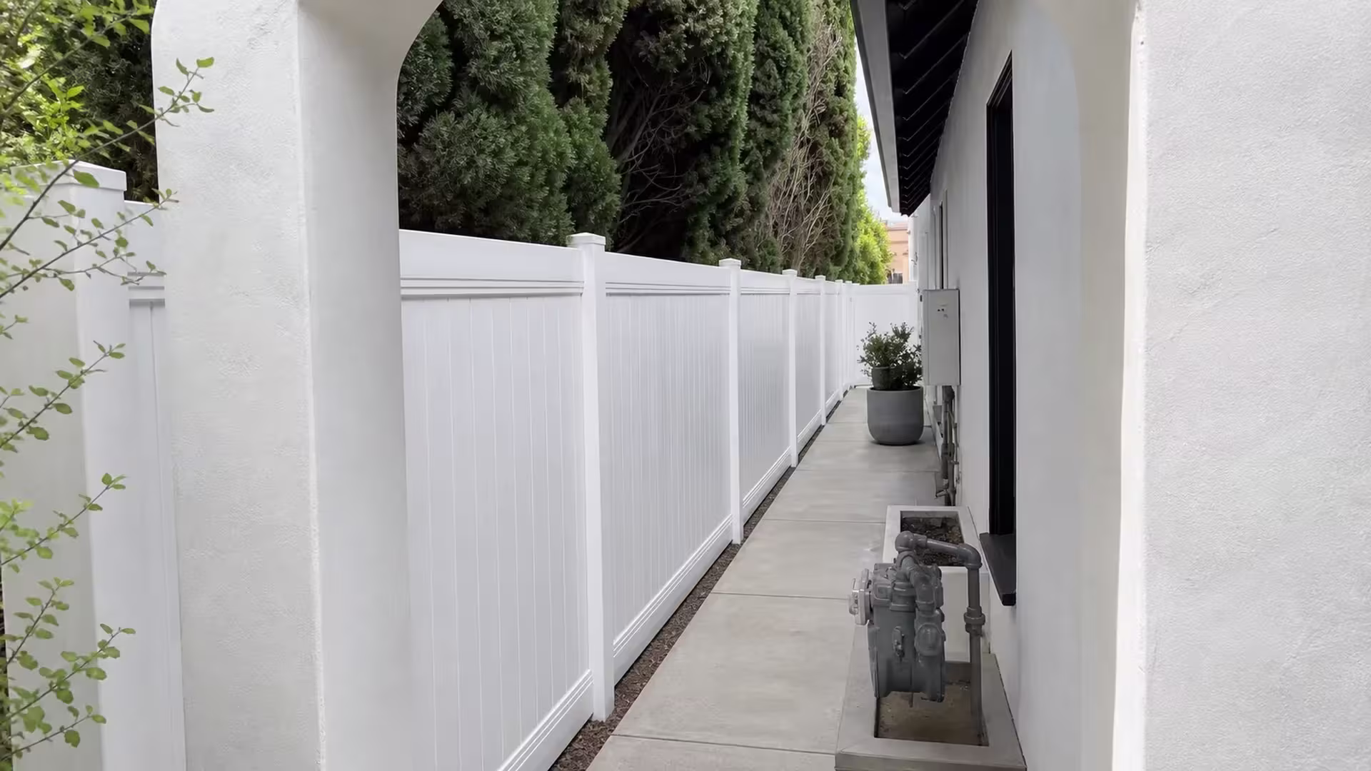 Dense hedges and utility pipes line this White vinyl fence and gate along a narrow Los Angeles walkway