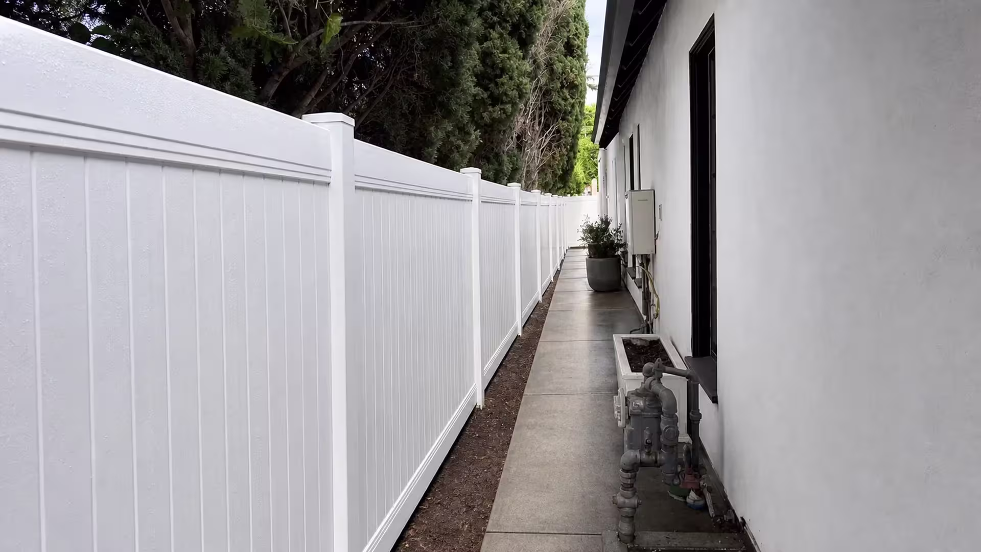A gas meter and regulator assembly sit beside this White vinyl privacy fence lining a Los Angeles walkway