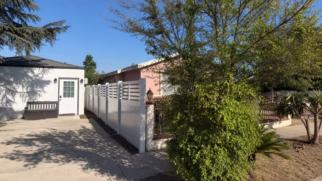 A horizontal picket top crowns an Aspen vinyl privacy fence angling past an outbuilding toward a pink neighboring building