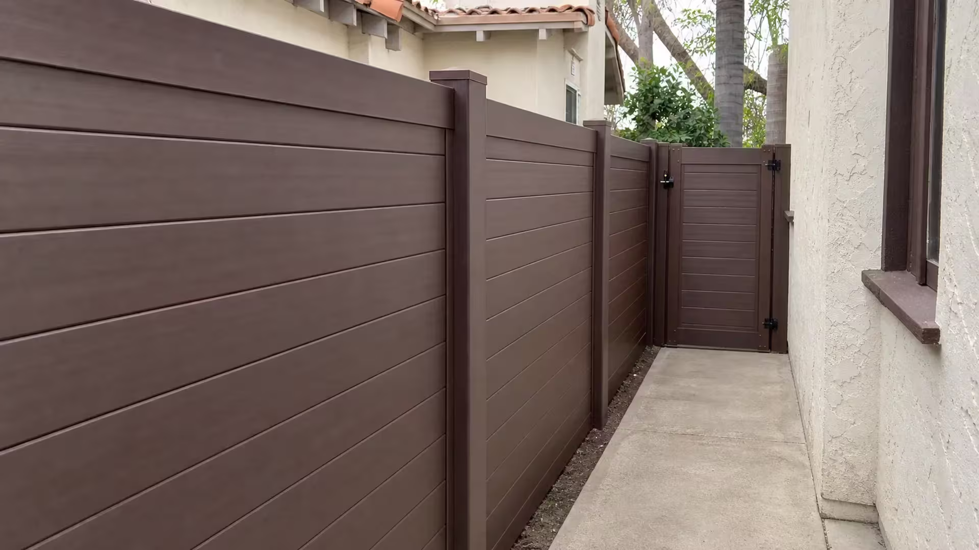 Clay tile roofline details frame this Dark Sequoia vinyl fence and gate beside a concrete walkway in Los Angeles