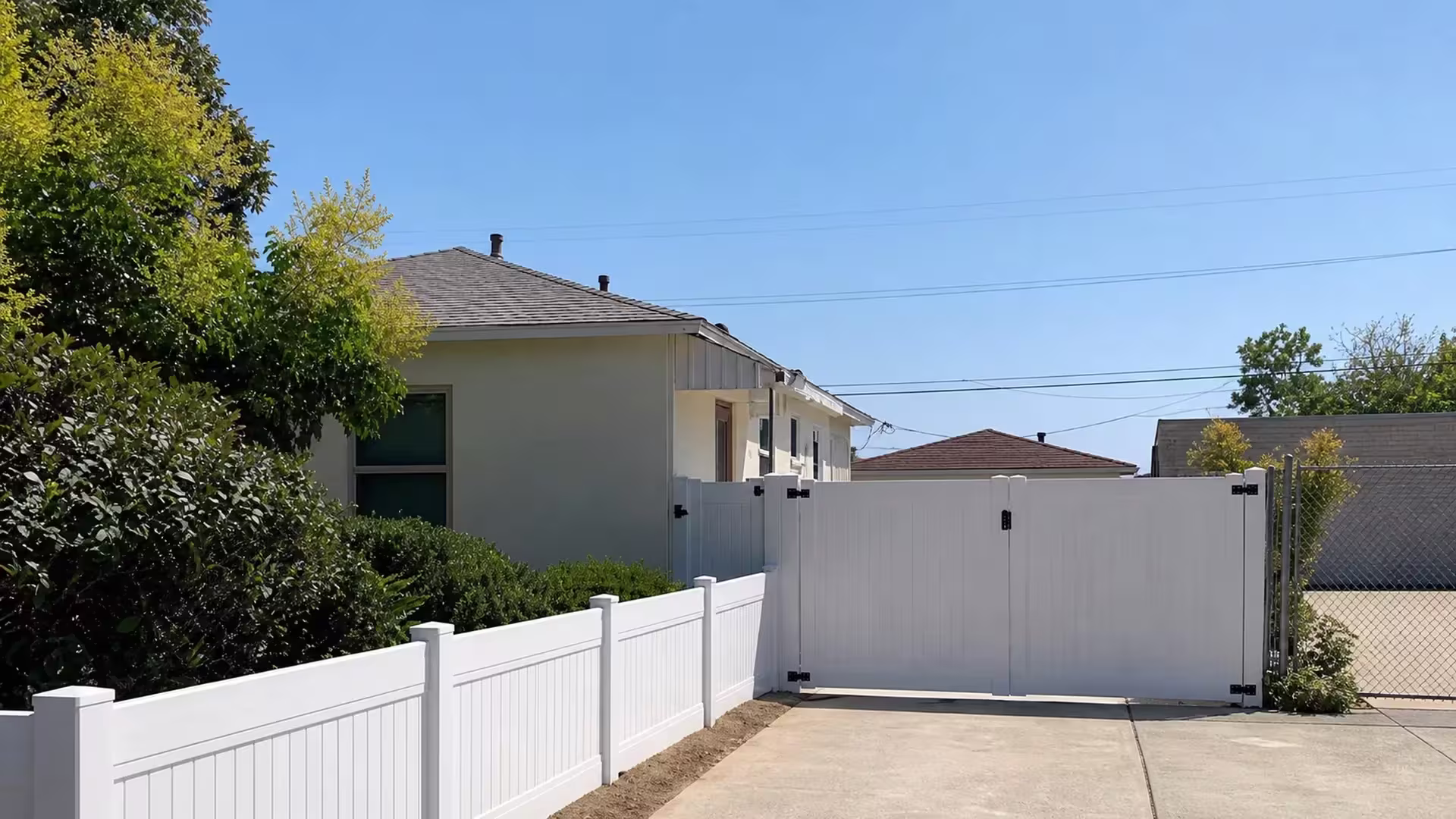 A beige house and a section of chain-link boundary flank this White vinyl privacy fence beside a paved driveway