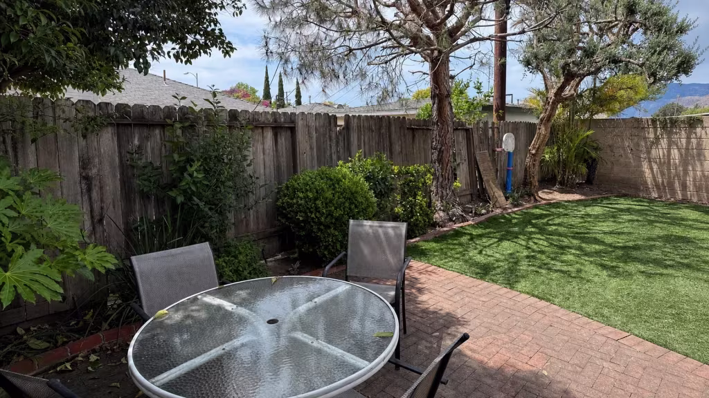 Before installation, a weathered wood fence borders a backyard with a brick patio, artificial grass, shrubs, and mature trees.