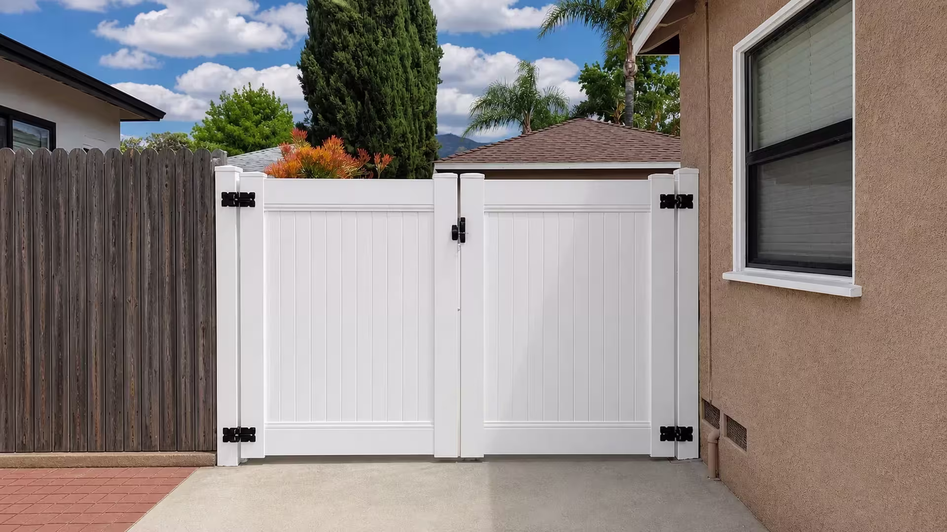 Brick-edged paving runs beside a White vinyl double privacy gate shaded by tall cypress trees in Shadow Hills