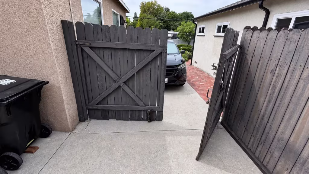 Before installation, open gray wood double gates span a driveway between two houses beside a trash bin and parked car.