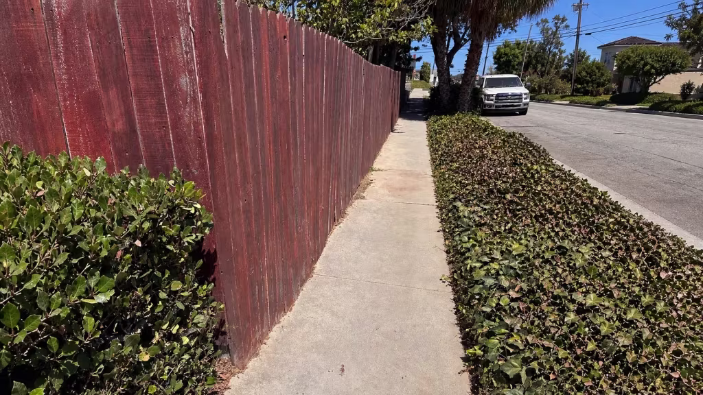Before installation, a faded red wood fence runs beside a public sidewalk and trimmed hedge.