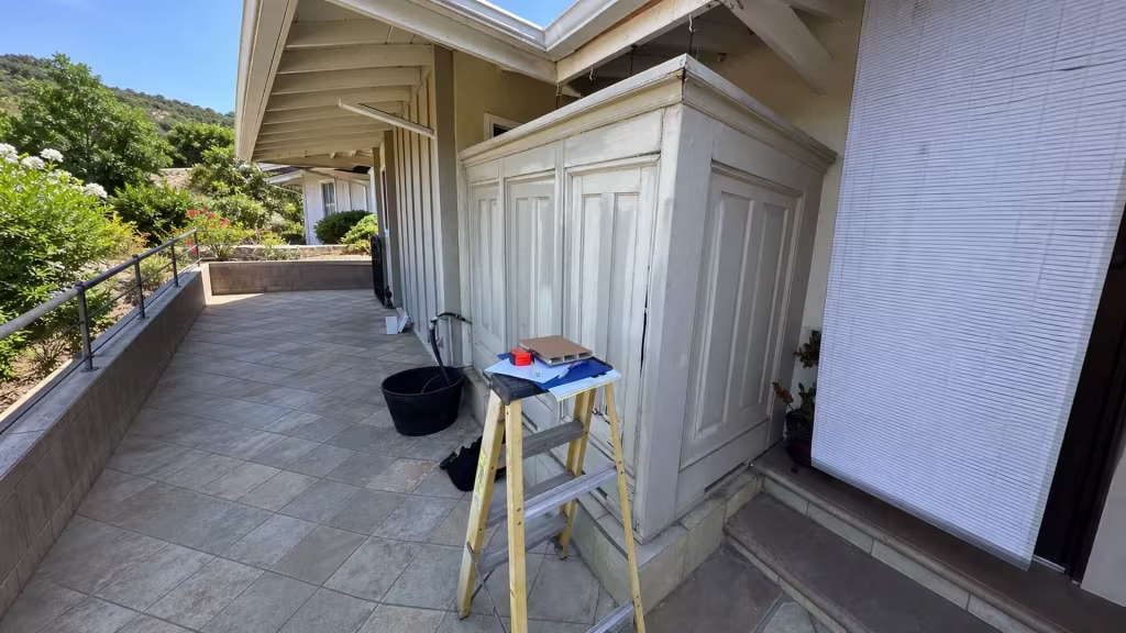 Before installation, a covered tiled patio contains an older white paneled enclosure, ladder, and low retaining wall.