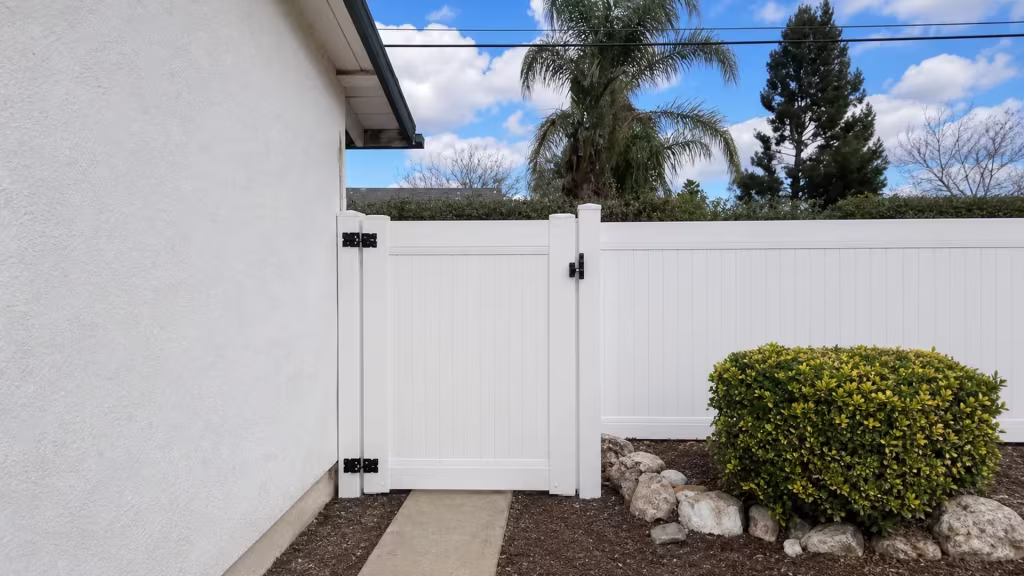 A trimmed shrub and landscape rocks line this West Hills passage, where a White vinyl gate closes the privacy fence