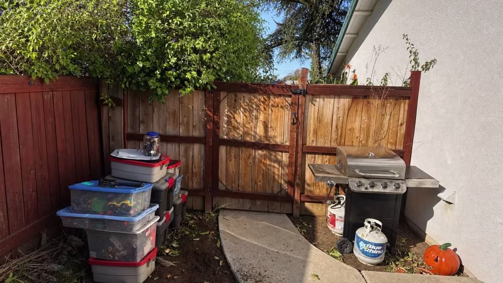 Before installation, a wood double gate closes a concrete path beside stacked storage bins, a grill, and leafy trees.