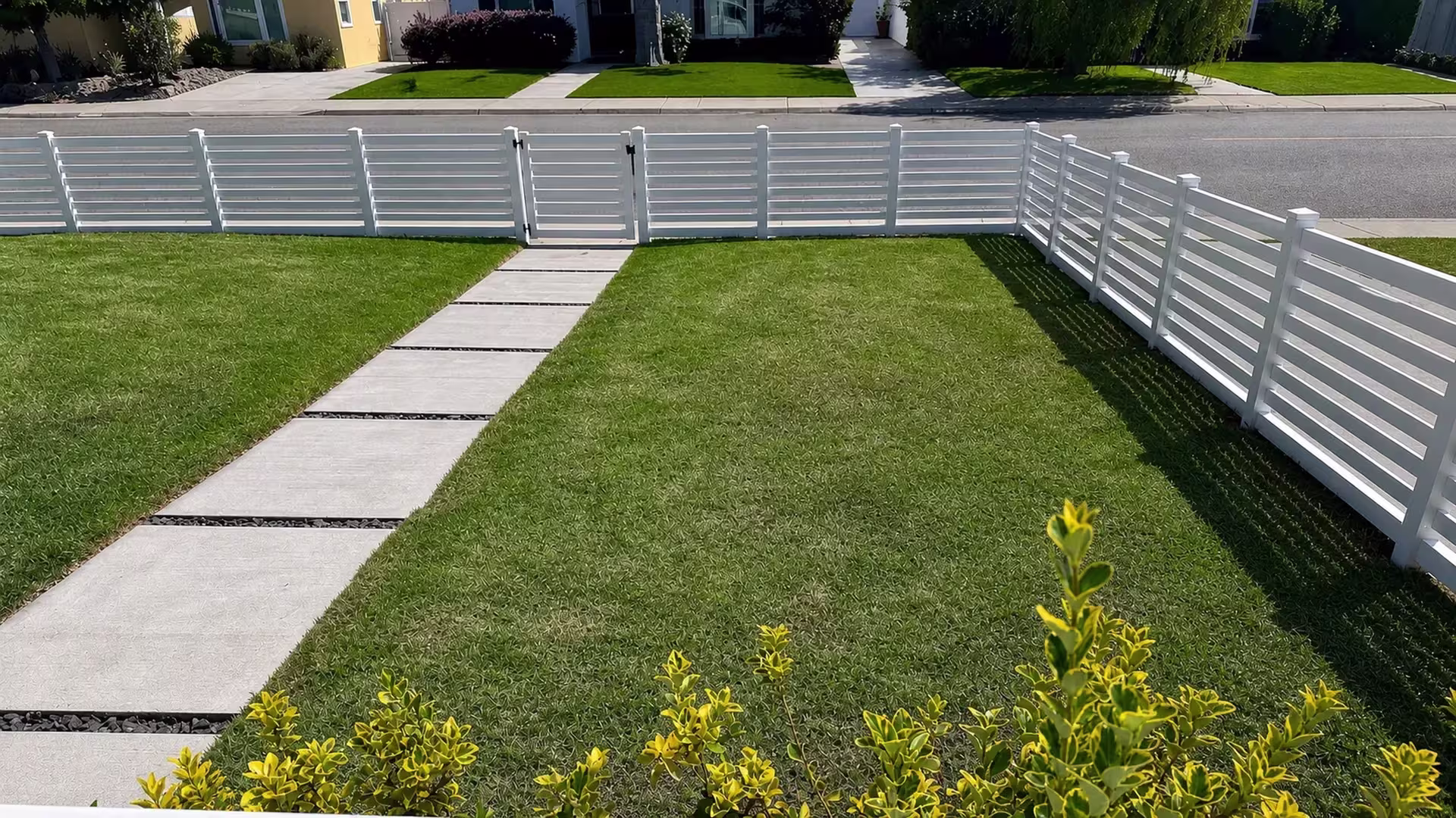 A White vinyl horizontal picket fence borders the open front lawn along a concrete walkway with dark stone joints