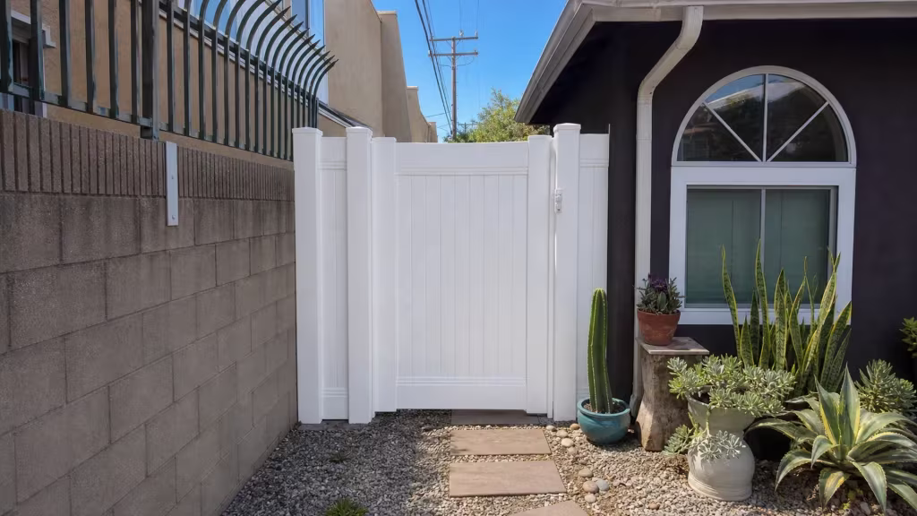 White vinyl picket fence gate set into a gray block wall beside a charcoal stucco house in Lakewood