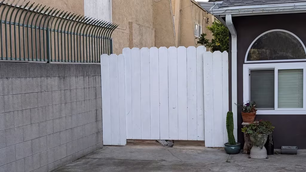 Before installation, a white-painted wood gate stands between a gray block wall and house beside several potted plants.