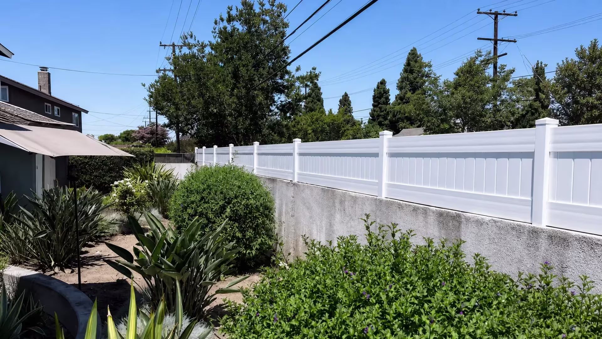 Succulents and flowering plants border the neighboring building beside a White vinyl privacy wall topper here