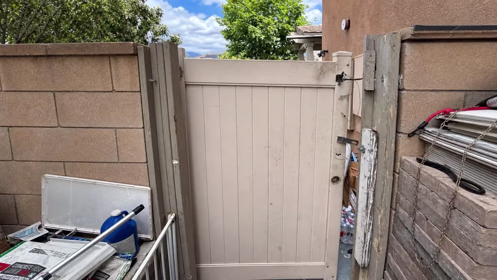 Before installation, a mismatched older gate panel stands between block walls amid stacked materials and stored items.
