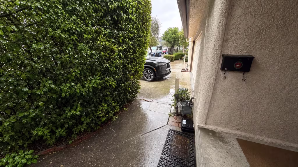 Before installation, a wet side walkway runs between a tall hedge and stucco house toward a parked vehicle.