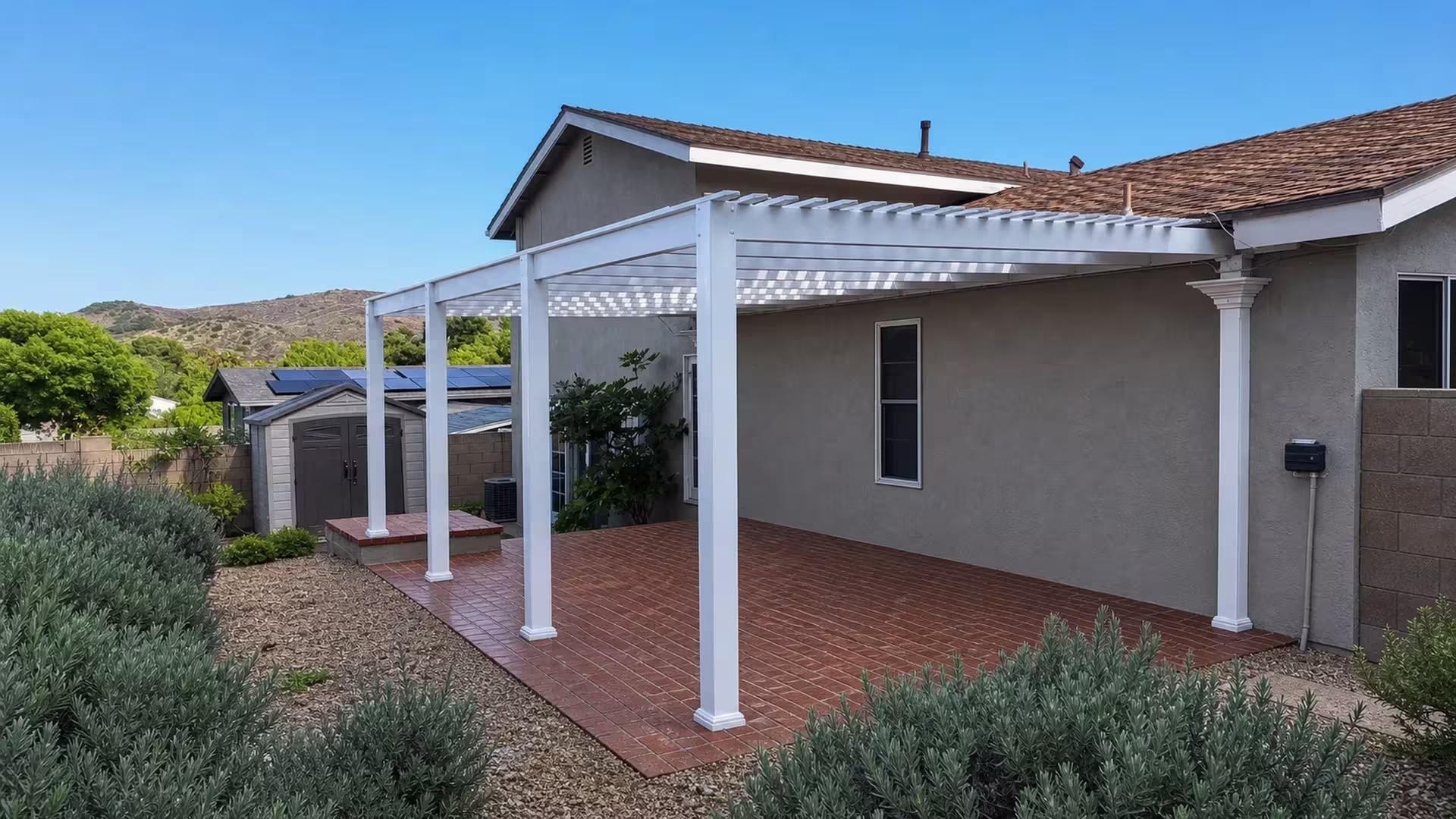 Rooftop solar panels show above the open lattice roof of this White vinyl pergola patio cover against a gray stucco wall
