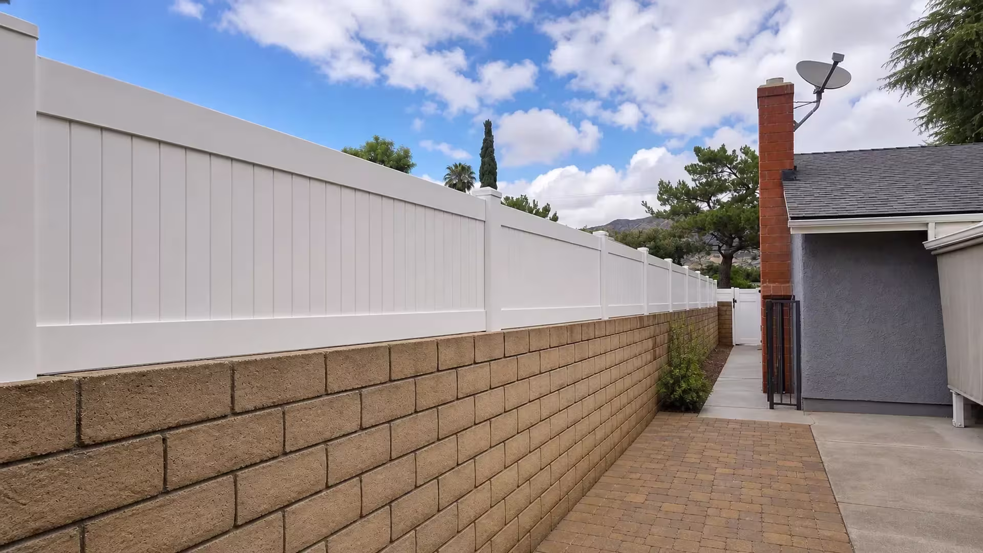 A brick chimney and satellite dish rise beyond this White vinyl privacy wall topper lining a Sunland yard