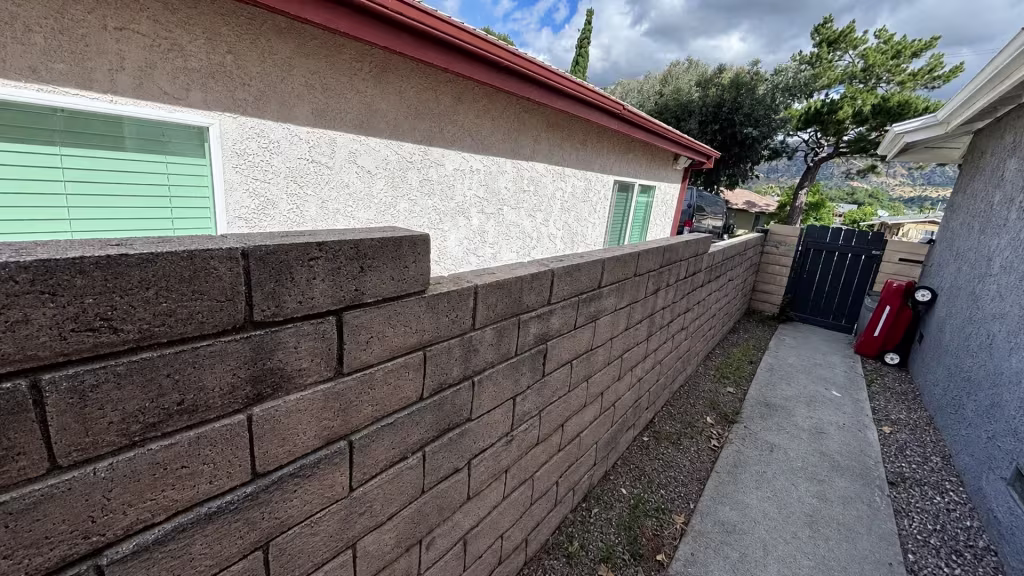 Before installation, a tan block wall borders a narrow walkway between houses and ends at a small dark gate.