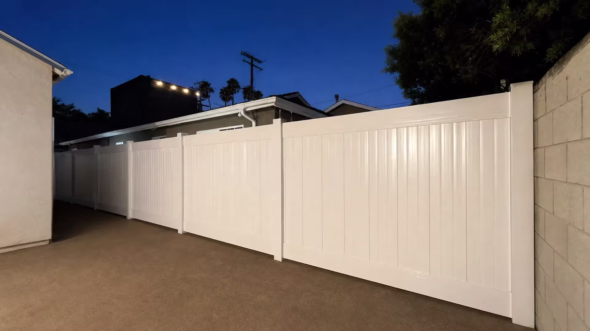 A pale masonry wall and a dark tree canopy stand where this White vinyl privacy fence ends in Culver City