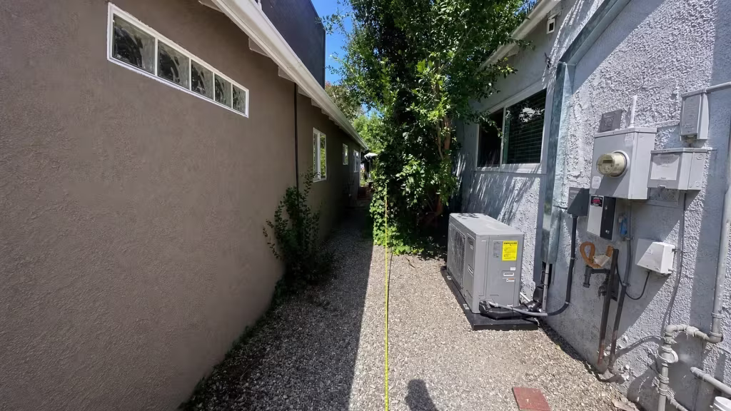 Before installation, a gravel side passage between two houses contains an air-conditioning unit and dense tree growth.