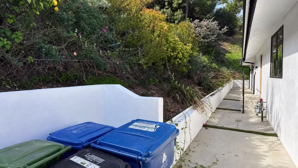 Before installation, a low white retaining wall follows a concrete path beside a densely landscaped slope and trash bins.