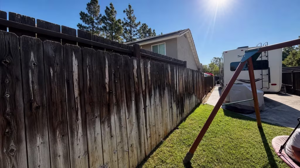 Before installation, a heavily weathered wood fence borders a grassy backyard beside a swing set and parked trailer.