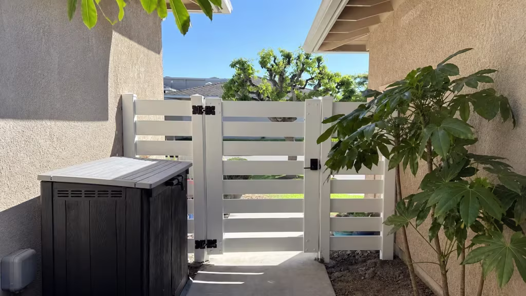 A narrow San Clemente passage opens to this White horizontal-picket vinyl gate and fence, past soil beds and tall foliage