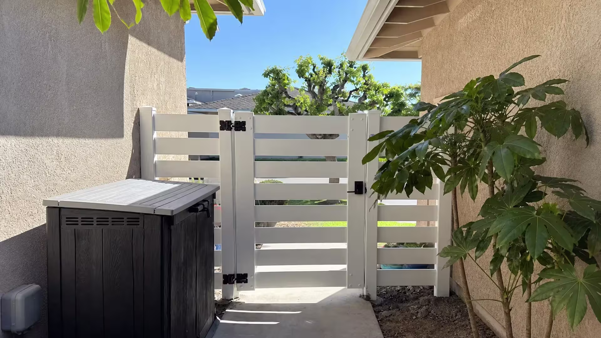 A narrow San Clemente passage opens to this White horizontal-picket vinyl gate and fence, past soil beds and tall foliage