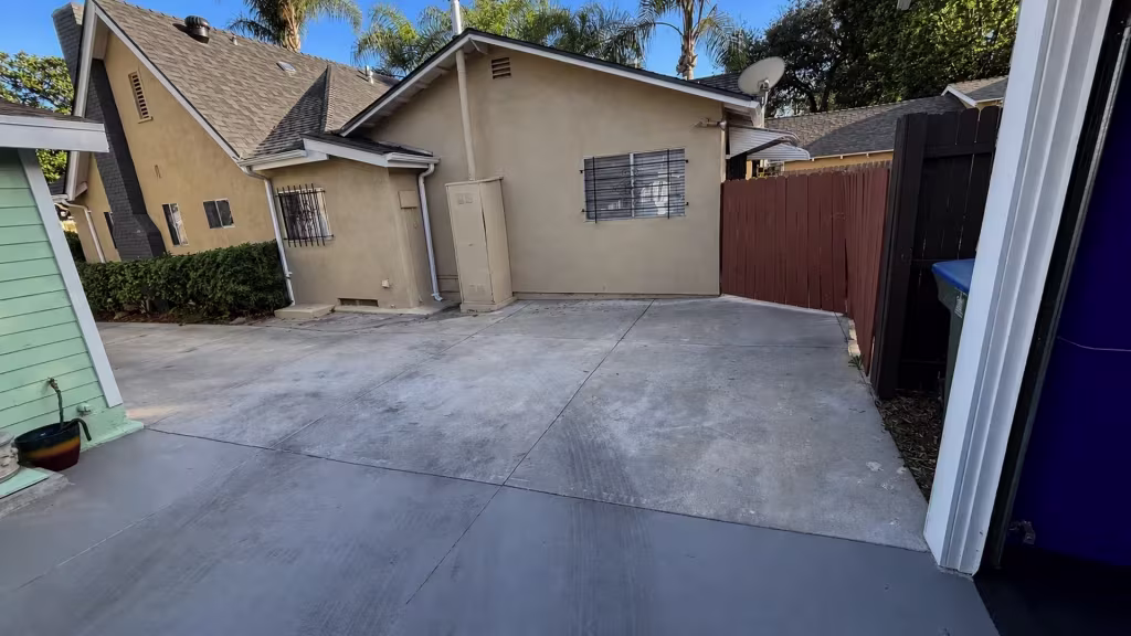 Before installation, an open concrete courtyard is bordered by a red-brown wood fence and neighboring stucco building.