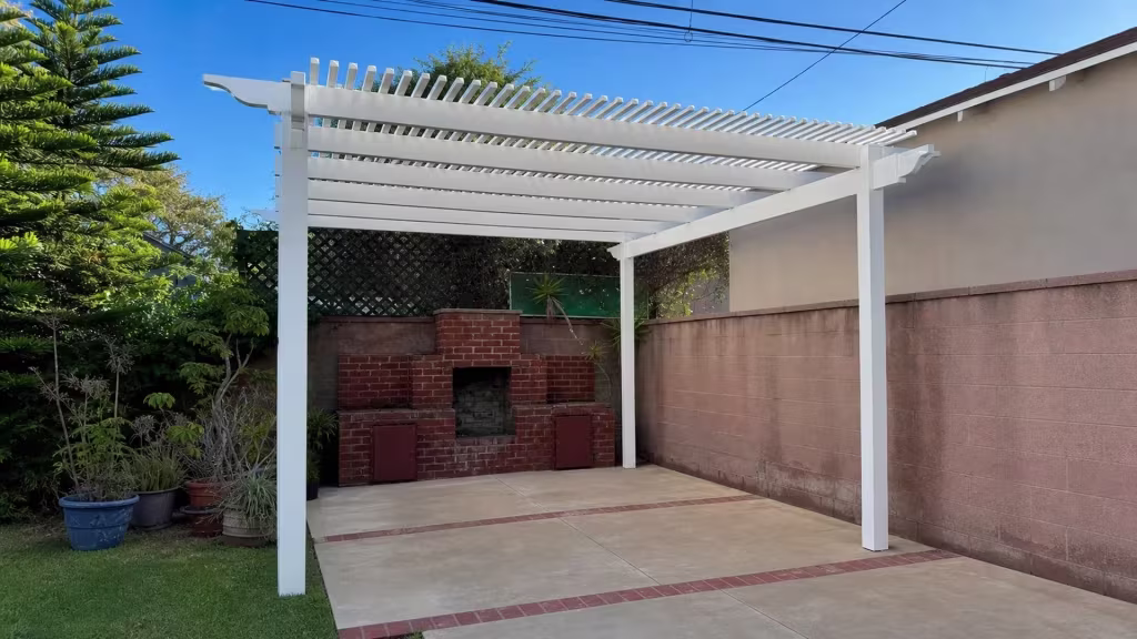 Evergreen trees and a pink concrete block wall frame this White manufactured-vinyl patio cover's open lattice roof