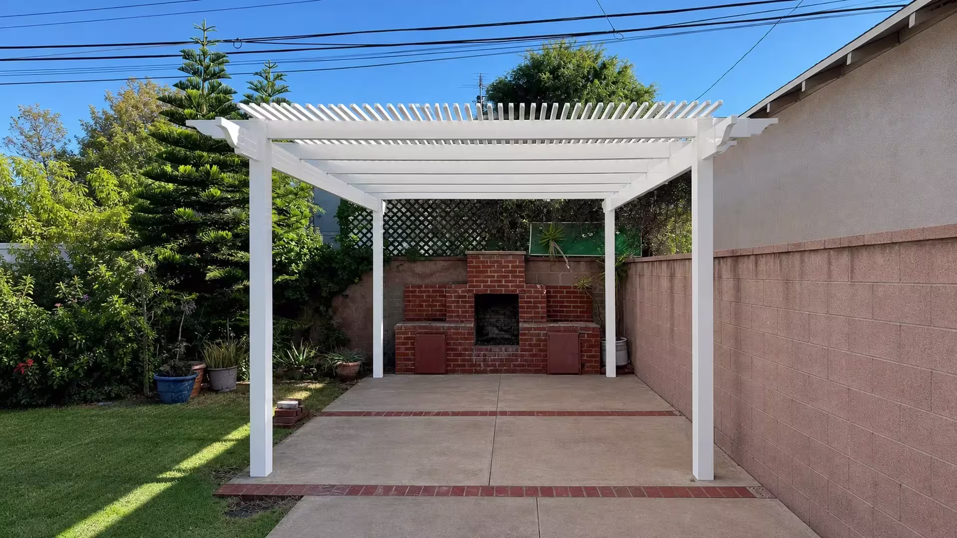 White vinyl open lattice patio cover with four square posts set beside a brick outdoor fireplace