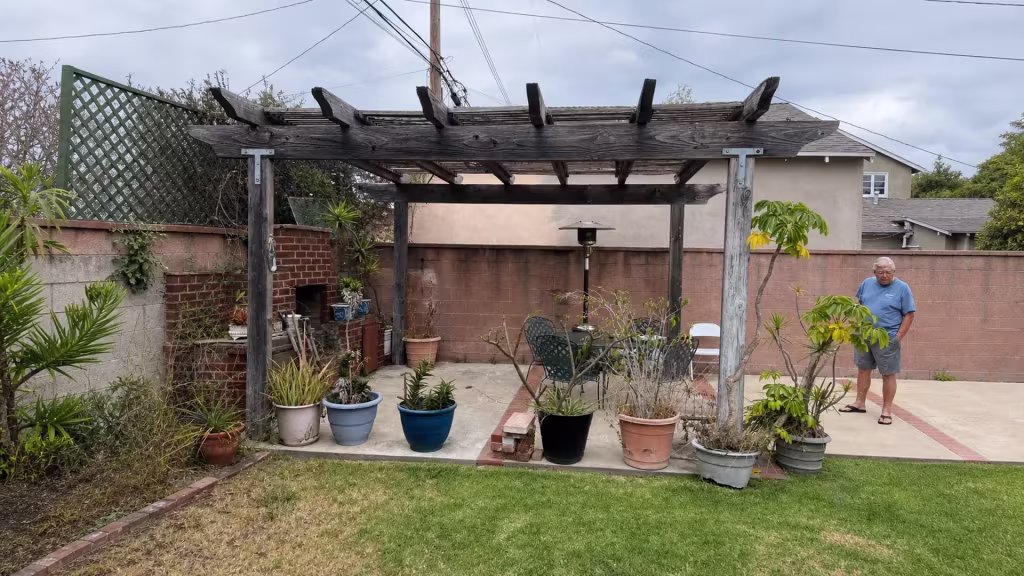 Before installation, an aging wood pergola shades a patio with potted plants beside brick and block walls.