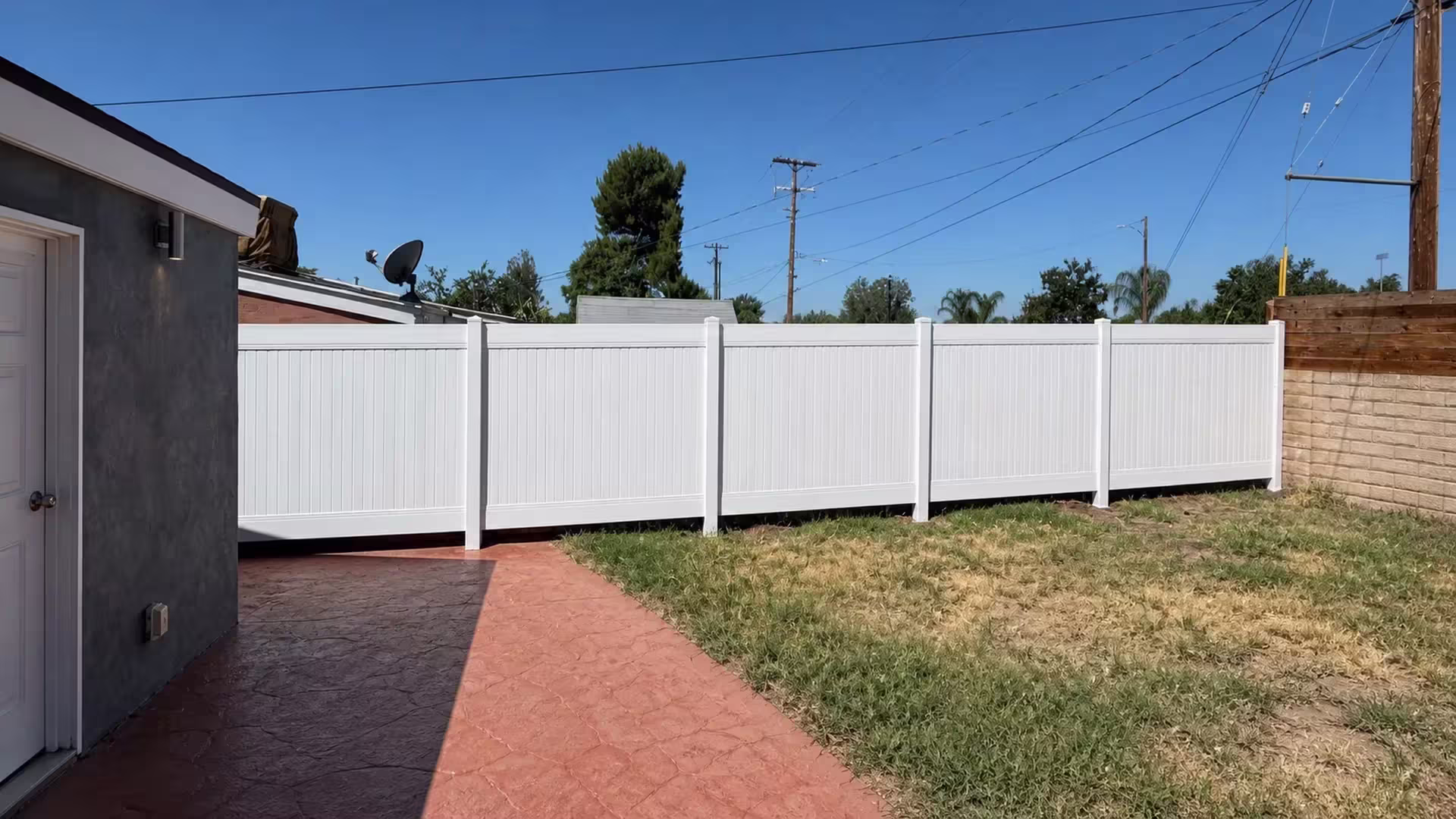 An exterior wall light and pale door trim mark the outbuilding beside this vinyl fence, finished in White, in Canyon Country