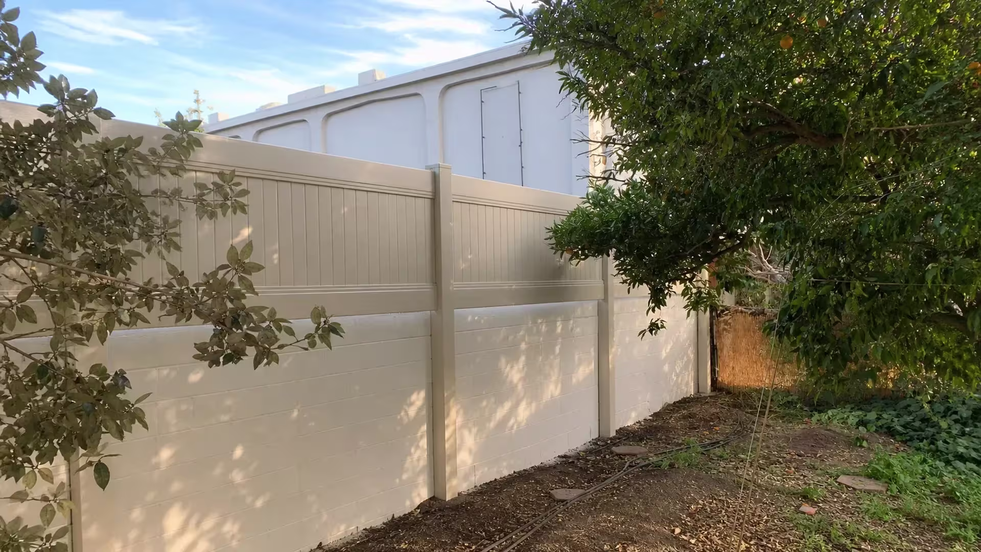 Tan vinyl fence panels continue over the Whittier block wall toward a shaded soil boundary