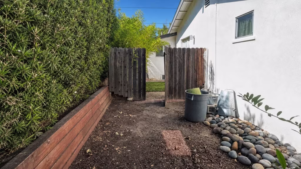 Before installation, open weathered wood gates lead into a side yard with a hedge, bare soil, river rocks, and a trash bin.