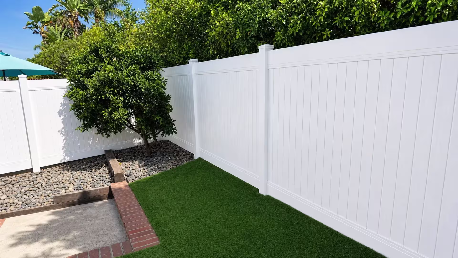 A turquoise patio umbrella stands near this White vinyl privacy fence turning past a mature citrus tree in Laguna Hills