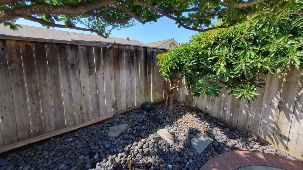 Before installation, a weathered wood fence surrounds a rock-covered garden corner beneath leafy trees.
