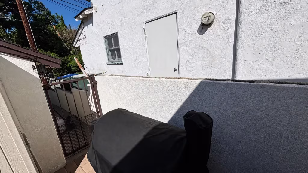 Before installation, a low white stucco divider and brown metal gate border a small patio beside the house.