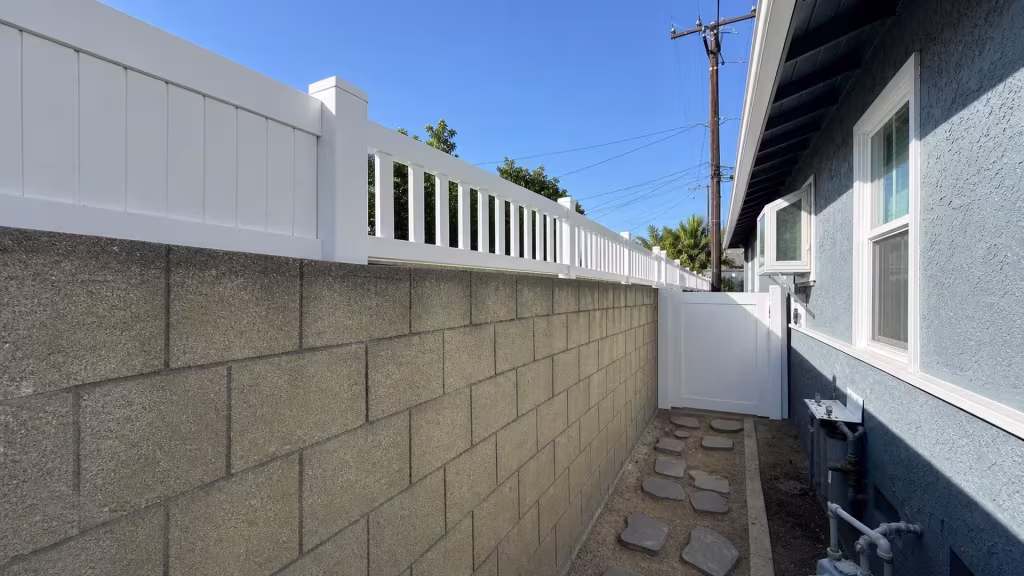 White vinyl wall-top privacy fence lines a gray masonry wall across from a blue-gray stucco house in Long Beach