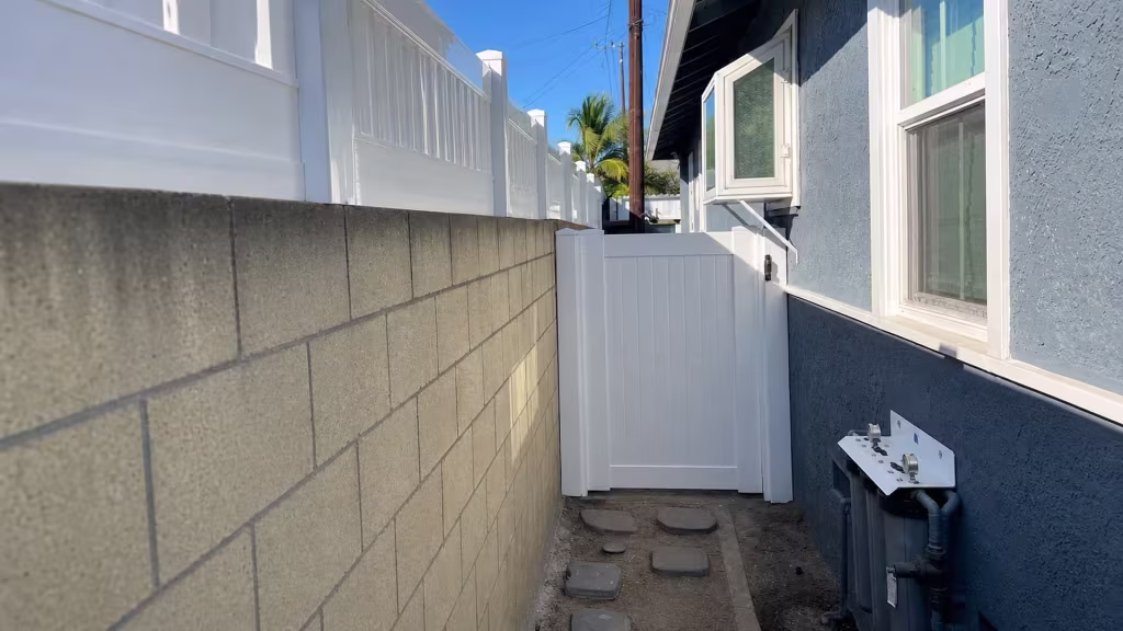 This White vinyl privacy fence's gate leaf closes a narrow dirt passage lined with gray stepping stones in Long Beach