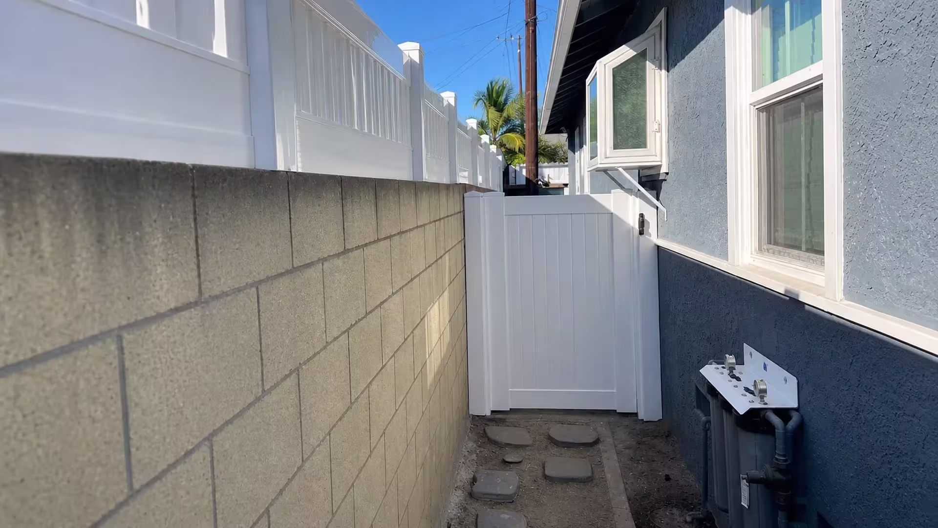 This White vinyl privacy fence's gate leaf closes a narrow dirt passage lined with gray stepping stones in Long Beach