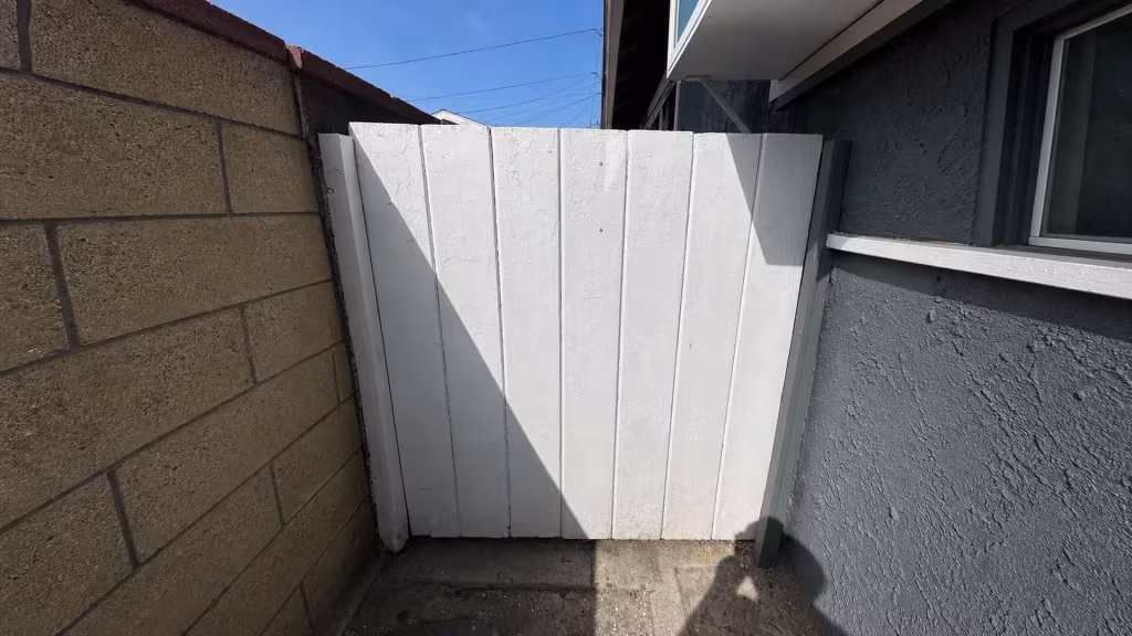 Before installation, a white wood gate closes a narrow passage between a tan block wall and gray stucco house.