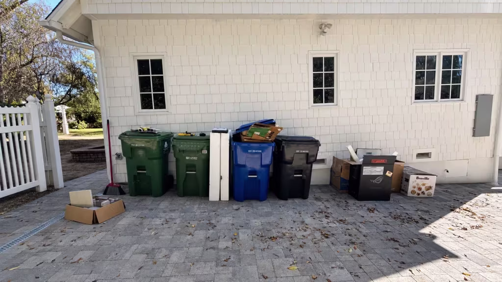 Before installation, a paved side yard beside a white house contains trash bins, cardboard boxes, and an open picket gate.