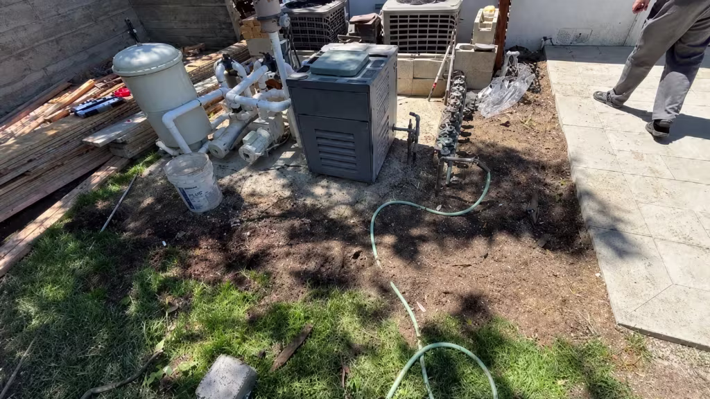 Before installation, a bare equipment area contains pool machinery, hoses, stacked lumber, and unfinished block posts beside a tiled walkway.
