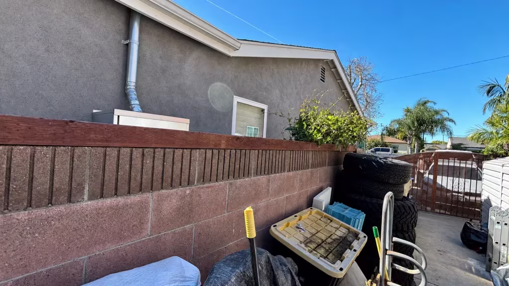 Before installation, a cluttered side yard contains stacked tires, bins, tools, a low block wall, and a rusty metal gate.