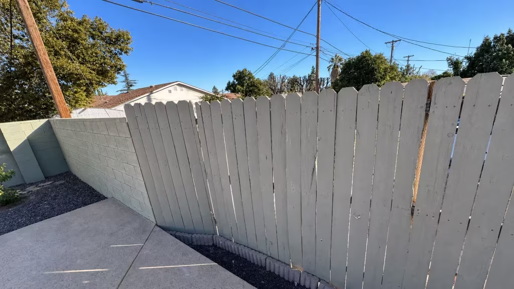 Before installation, a white-painted wood fence and gate border a concrete patio beside a pale block wall.