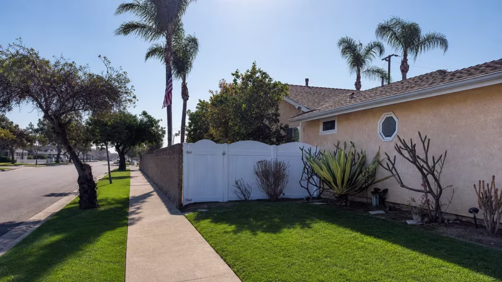 White vinyl privacy fence lines a Huntington Beach sidewalk, with a curb and grass strip framing the street edge