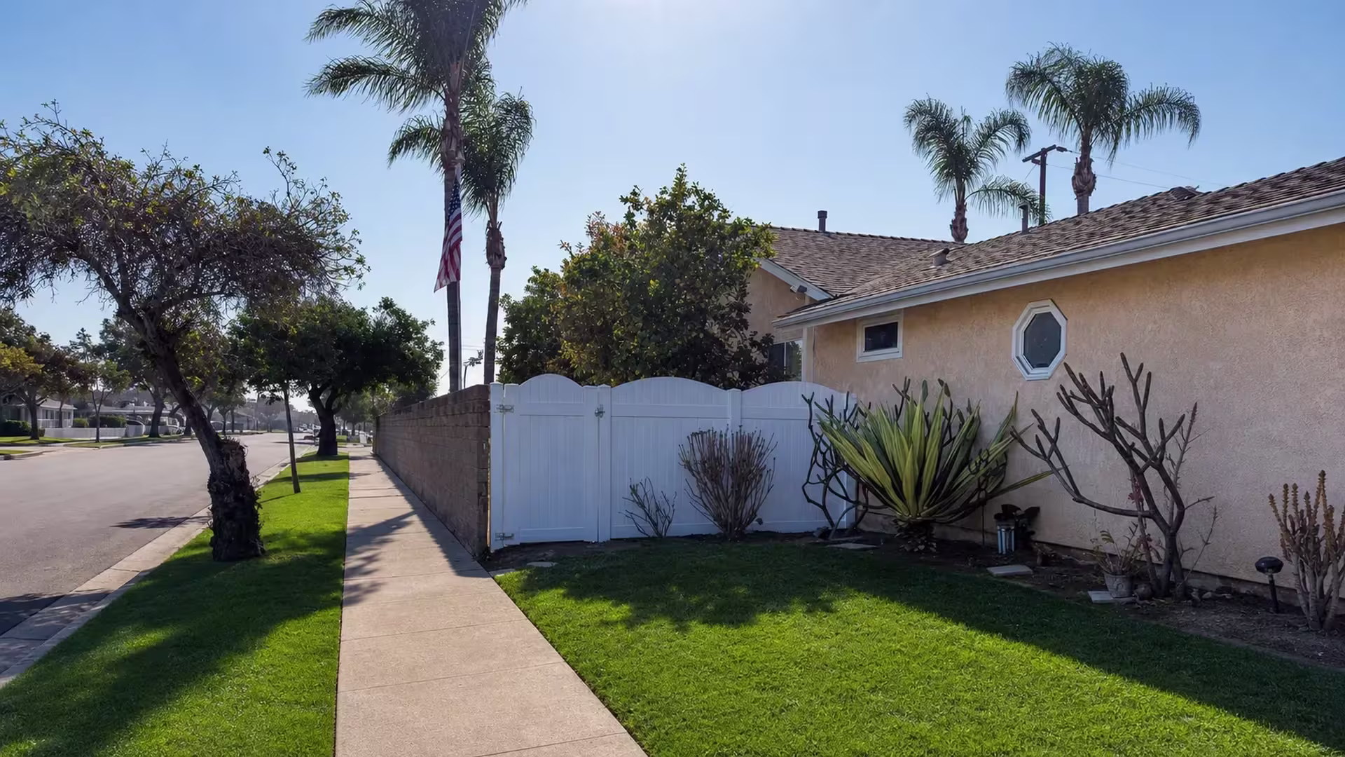 White vinyl privacy fence lines a Huntington Beach sidewalk, with a curb and grass strip framing the street edge