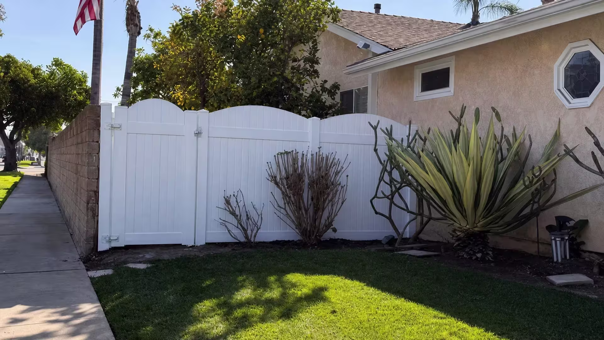 White vinyl privacy fence gate stands beside a tan masonry boundary wall near a sculptural agave planting in Huntington Beach