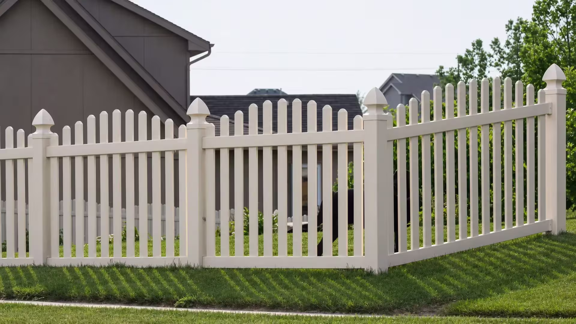Tan vinyl arched picket fence lining a sloping front lawn beside neighboring suburban houses