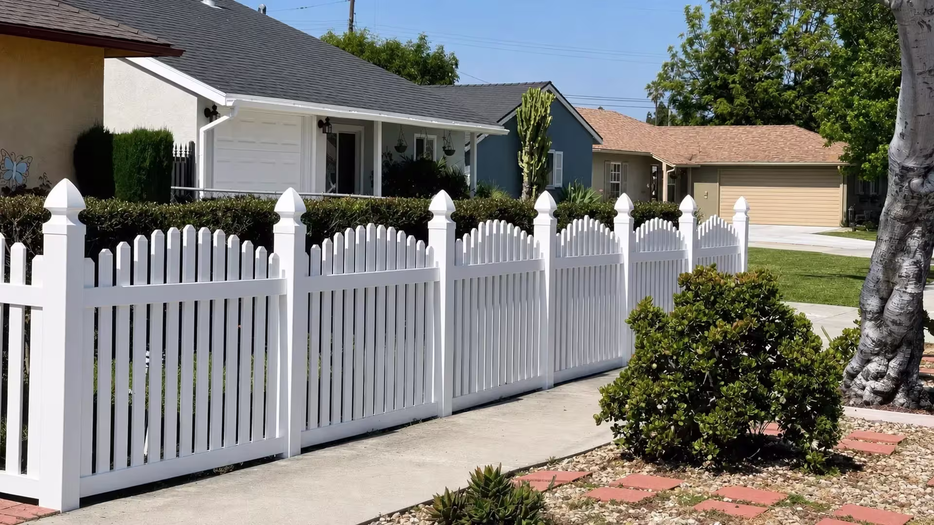 Cactus and gravel replace lawn along the driveway edge traced by White vinyl arched picket fence
